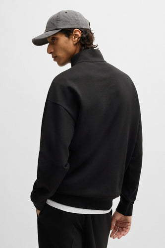 Man met grijze pet, zwarte trui en broek op een witte achtergrond.