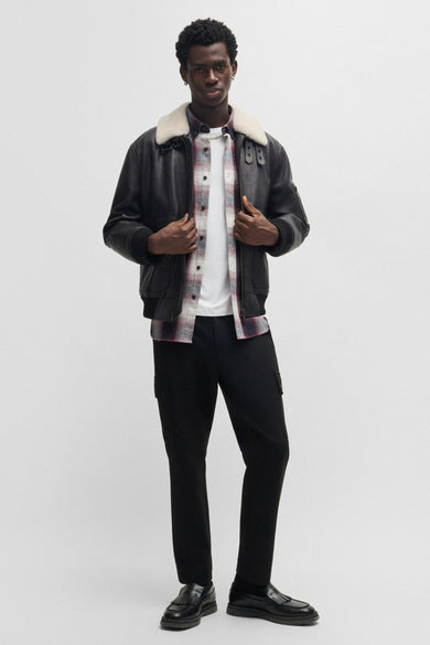 Man in zwarte jas met lamswollen kraag, geruite blouse, witte T-shirt en zwarte broek.