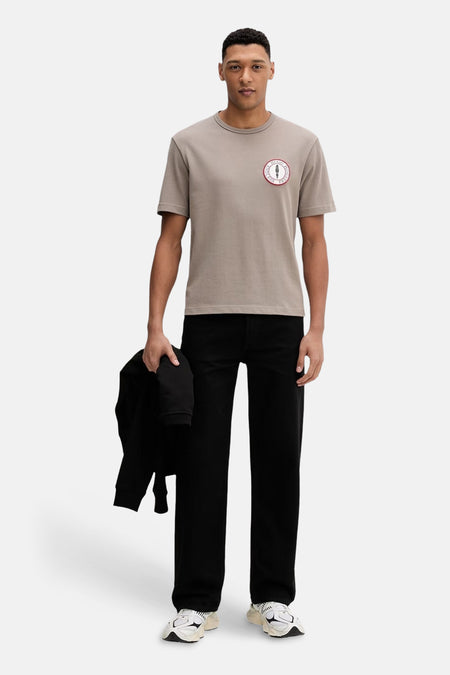 T-shirt marron de HUGO, avec un badge rond sur la poitrine et porté avec un pantalon noir et des baskets.