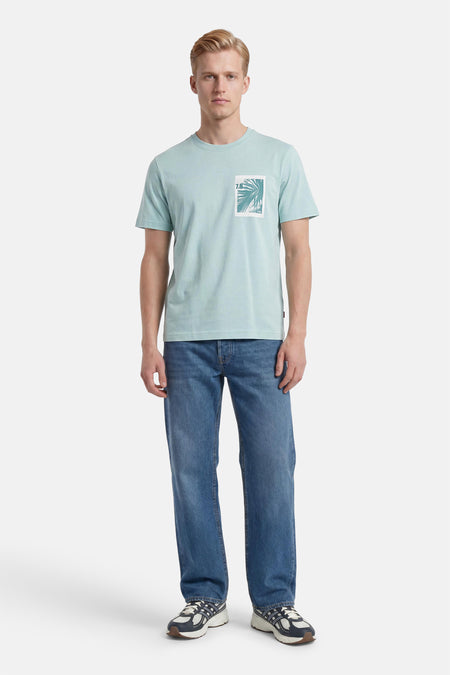 T-shirt turquoise à manches courtes de HUGO BOSS ORANGE, avec imprimé feuilles de palmier et '7.8' sur la poitrine, porté avec un jean bleu et des baskets.
