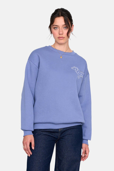 Pull-over bleu avec col rond de 24 colours gmbh, brodé d'un chien, porté avec des jeans foncés et une chaîne en or.