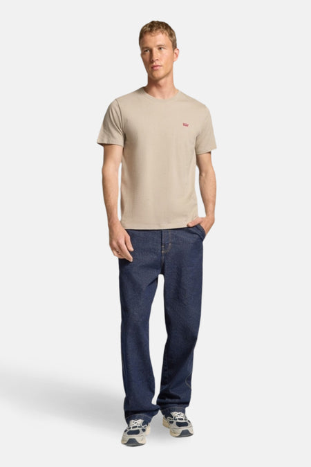 Een man draagt een beige T-shirt en blauwe spijkerbroek.