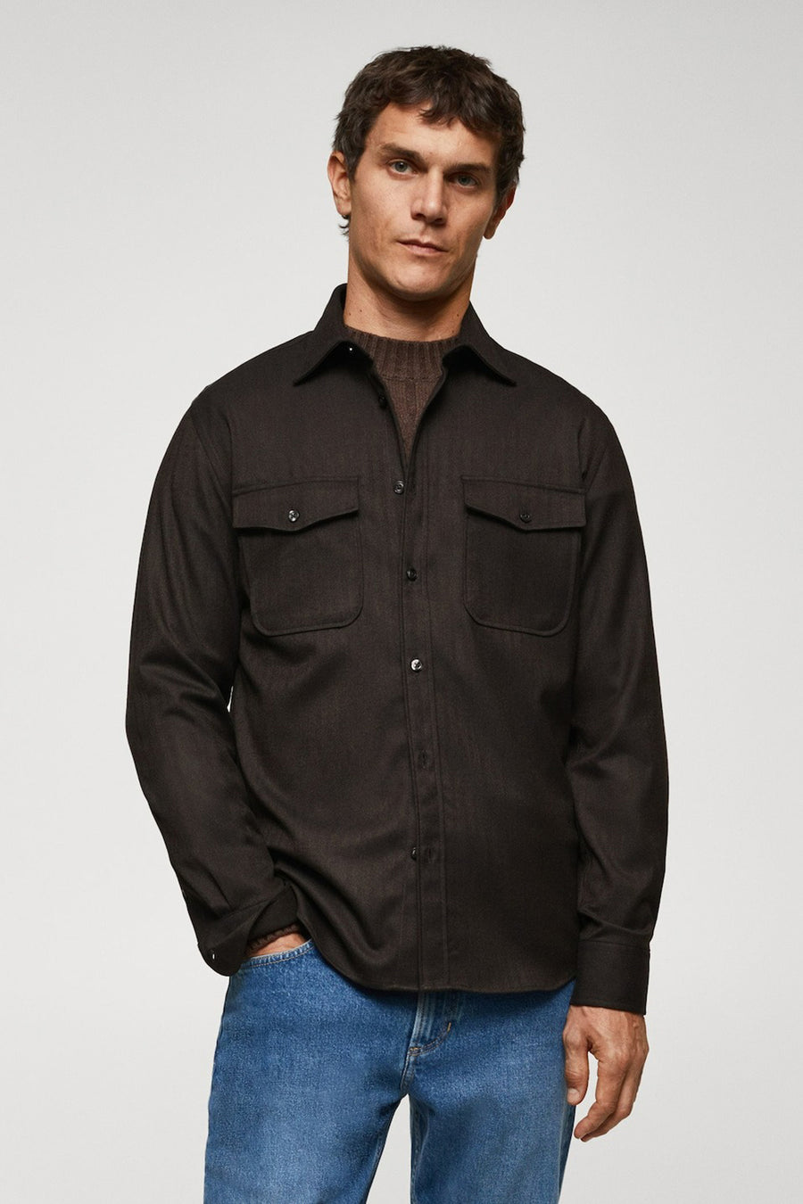 Chemise brune à manches longues de MANGO, avec poches po poitrine et portée sur une veste brune à col rond et des jeans bleus.