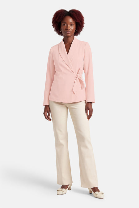 Roze blazer van Terra di Siena, met lange mouwen en een strikceintuur, gecombineerd met crèmekleurige broek en schoenen.
