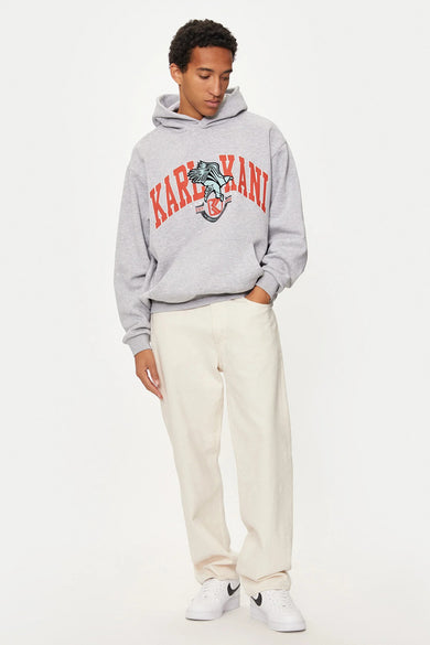 Pull-over gris avec capuche de Karl Kani, avec un logo sur la poitrine, associé à une légère pantalon et des baskets blanches.