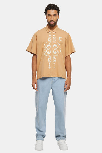 Chemise beige à manches courtes de Karl Kani, avec une fermeture éclair et des motifs blancs décoratifs.