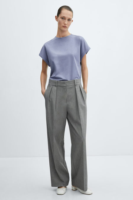 T-shirt bleu à manches courtes de MANGO, stylisé avec un pantalon gris plissé et des mules blanches.