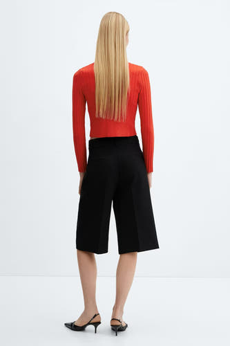 Gilet rouge de MANGO, avec des manches longues et un fin motif côtelé, porté avec des culottes noires et des escarpins élégants.