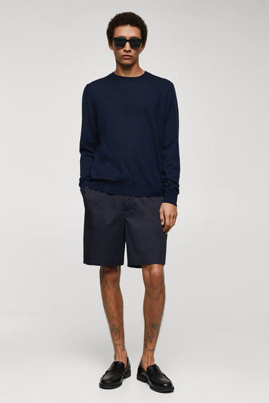 Short bleu de MANGO porté avec une chemise bleue marine et des loafers en cuir noirs, debout.