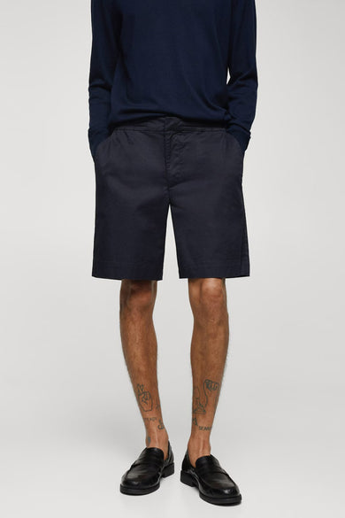 Short bleu de MANGO, porté avec une chemise bleue marine et des loafers noirs, avec des tatouages sur les coudes.