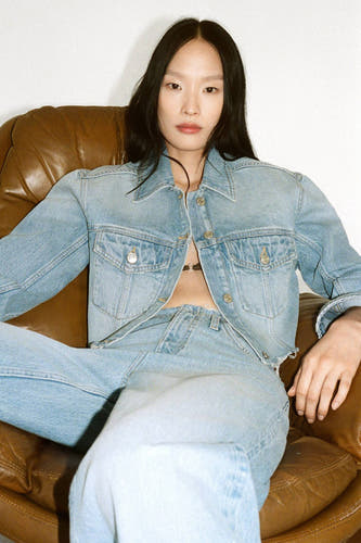 Spijkerjas in middenblauw denim van MANGO, gedragen met een zwarte bh en wijde jeans in een casual zithouding.
