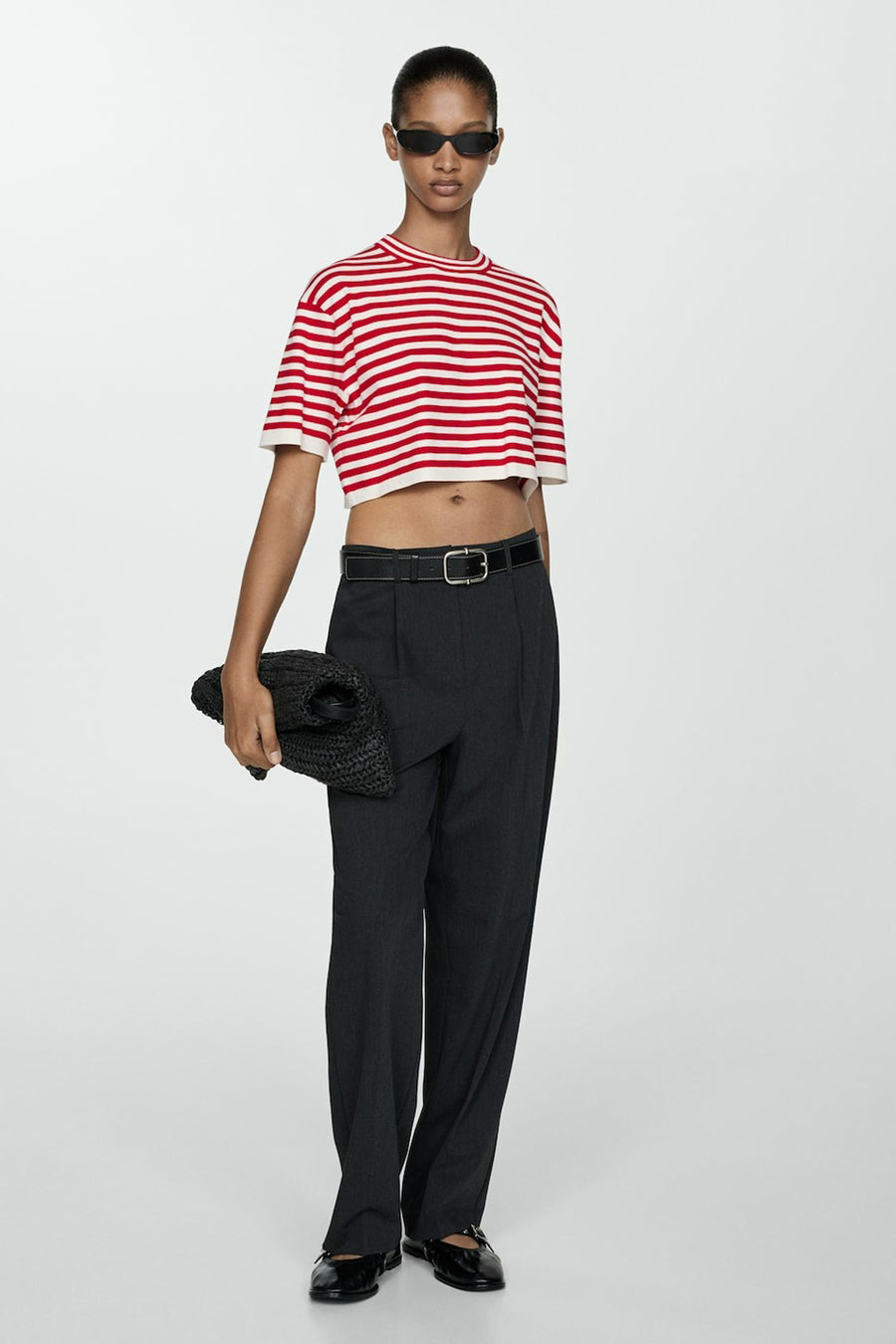 Een model staat met een gestreept shirt, een zwarte broek, een zwarte riem, zwarte schoenen en een zwarte tas.