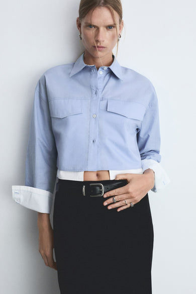 Chemise bleue à manches longues de MANGO, dos avec des manches roulées et pantalon noir avec ceinture.
