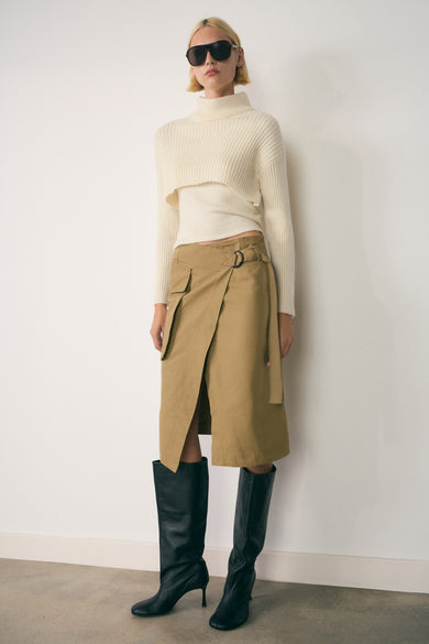 Veste blanche de MANGO, superposée à une veste en tricot à manches longues coupées, associée à un châle kaki et des bottes noires à talons.