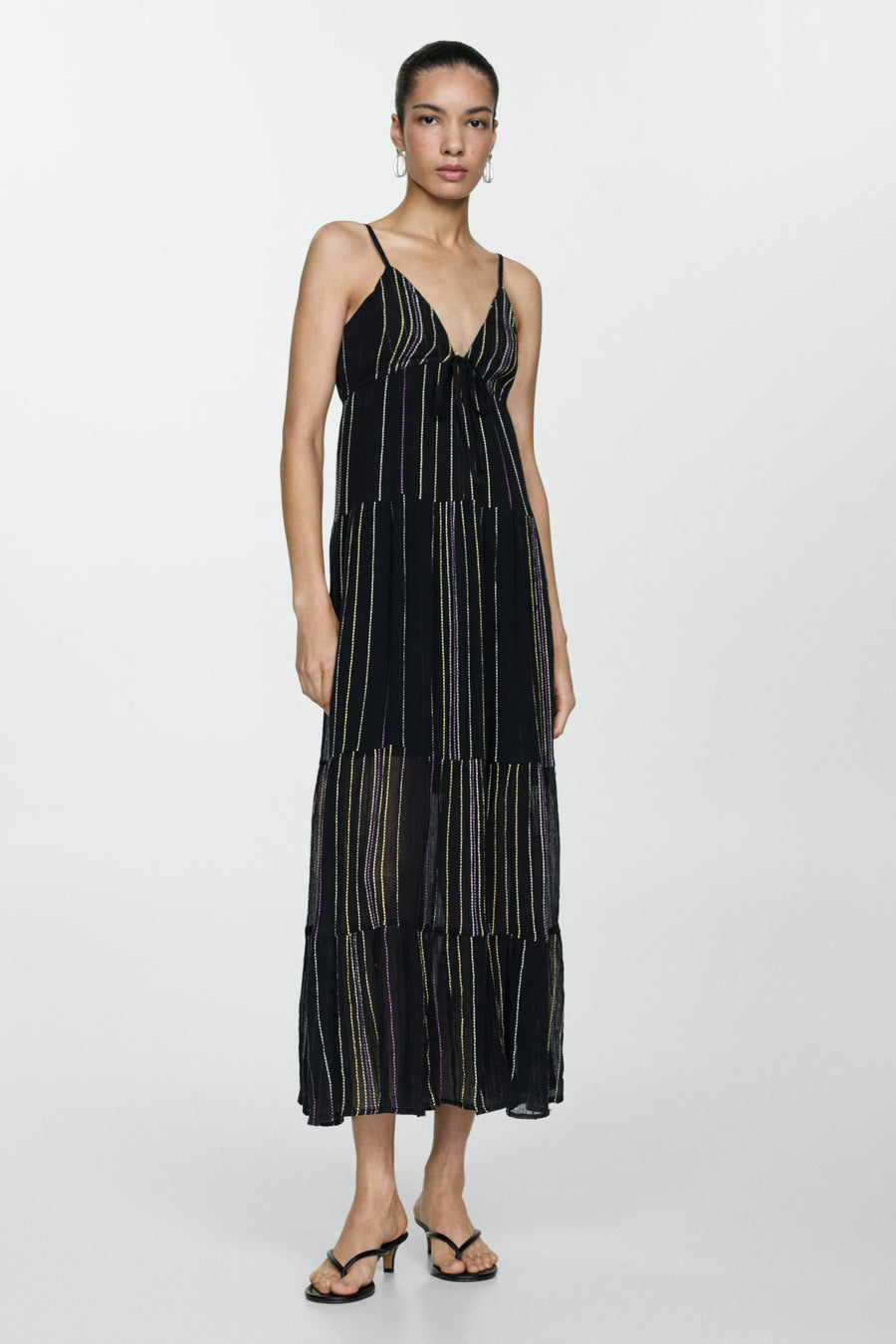 Vrouw in een lange, gestreepte, zwarte zomerjurk met spaghettibandjes en zwarte sandalen, staand tegen een witte achtergrond.
