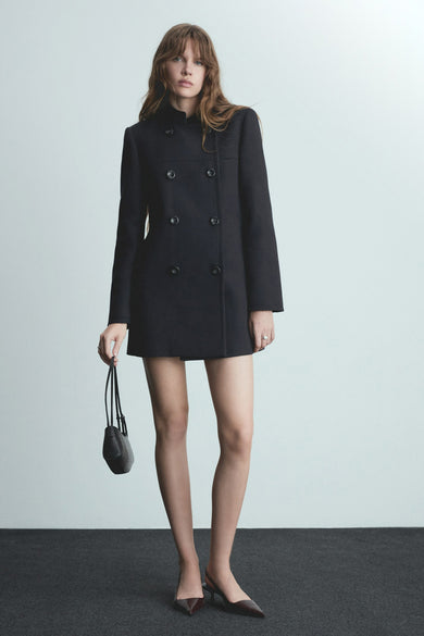 Manteau long bleu de MANGO, avec une double rangée de boutons, porté avec des bottes et une petite poche.