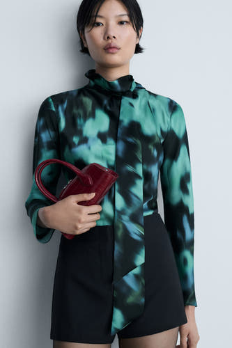Blouse multicolore à longues manches de MANGO, avec un motif abstrait et portée avec des shorts noirs et une pochette rouge.