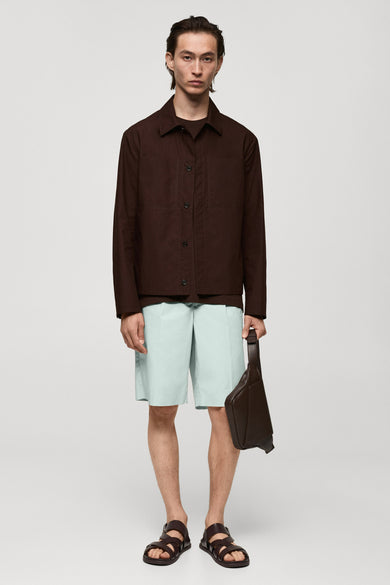 Manteau court rouge de MANGO, avec des manches longues, porté sur un T-shirt et des shorts légers avec une sacoche en cuir et des sandales.