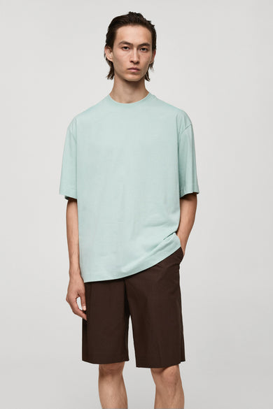 T-shirt bleu à manches courtes de MANGO, porté avec des shorts bruns, devant un fond uni.