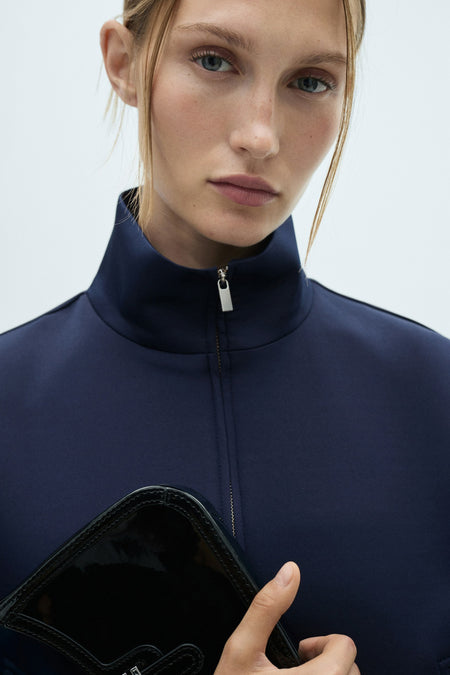 Gilet bleu de MANGO, avec un col haut et une sac noir dans la main.
