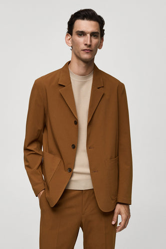 Oranje blazer van MANGO, met knoopsluiting en opgestikte zakken, gedragen over een crèmekleurige trui en bijpassende broek.
