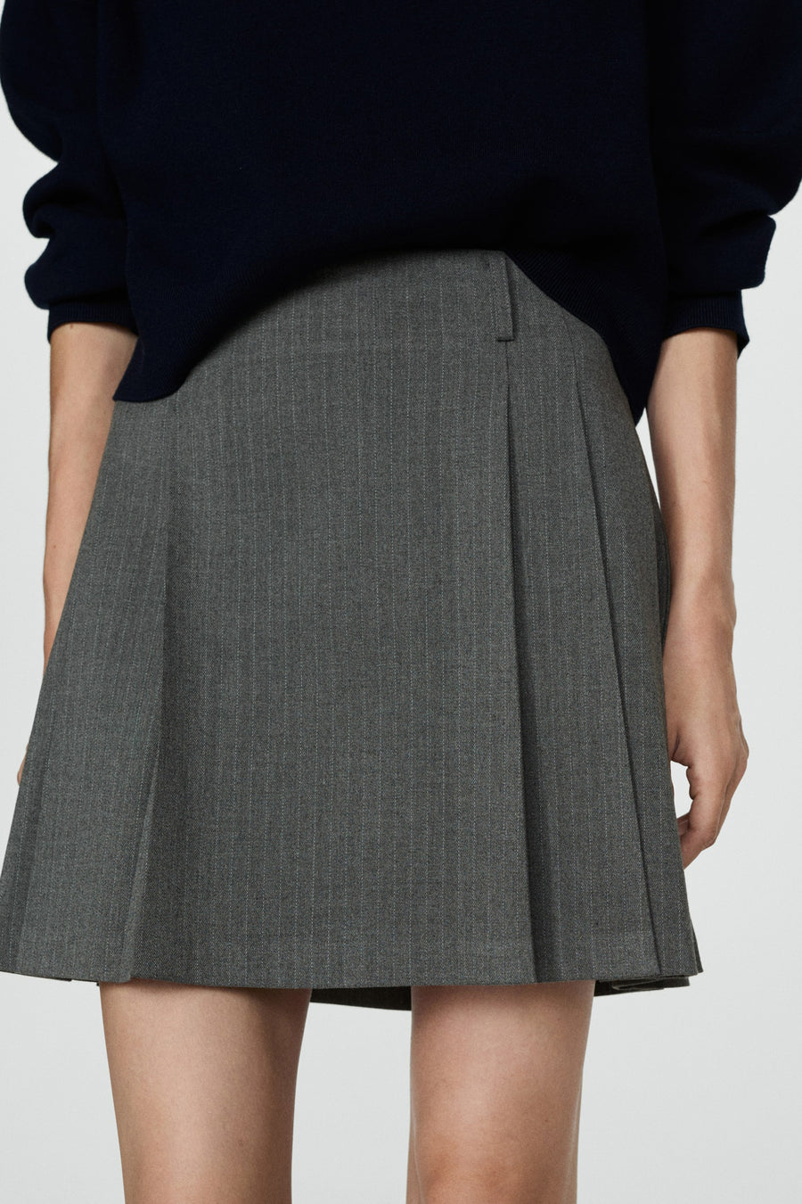 Jupe grise courte de MANGO avec une rayure et des plis, portée avec un pull bleu foncé.