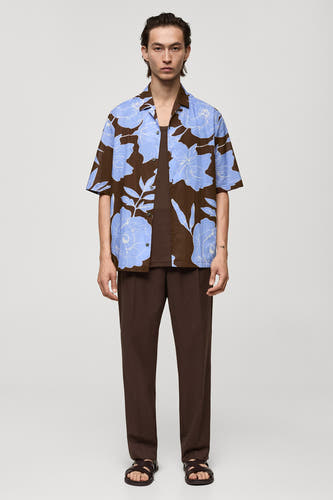 Chemise brune à manches courtes de MANGO, avec un motif de fleurs bleues, associée à des pantalons bruns et des sandales.