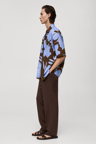 Chemise brune à manches courtes de MANGO, avec un motif de fleurs bleues, associée à des pantalons bruns et des sandales.