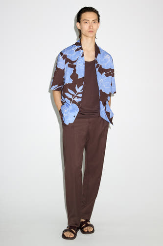 Chemise brune à manches courtes de MANGO, avec un motif de fleurs bleues, portée sur une chemise brune et un pantalon brune avec des sandales.