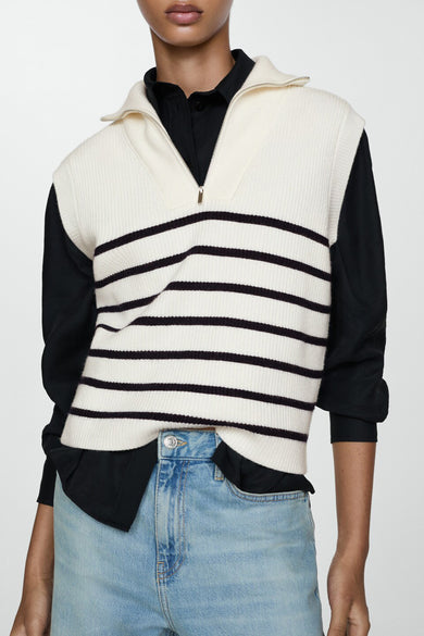 Débardeur beige de MANGO, avec des rayures noires horizontales et une fermeture éclair argentée, stylé avec une blouse noire et un jeans bleu.