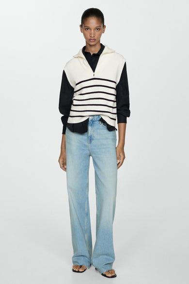 T-shirt beige de MANGO, avec des rayures sombres et une courte fermeture éclair, porté sur un chemisier noir à manches longues et un jean bleu.