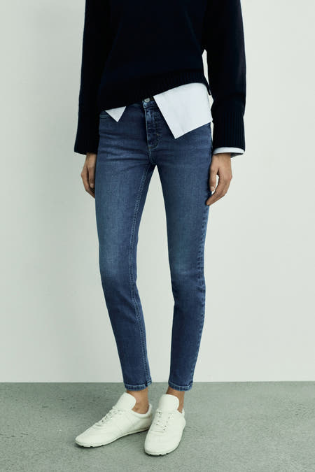 Jean skinny en denim bleu moyen de MANGO, associé à des baskets blanches et un pull noir.