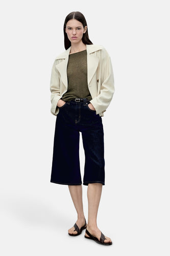 Vrouw met lichte trenchcoat, olijfgroene top, donkerblauwe culottes en leren sandalen.
