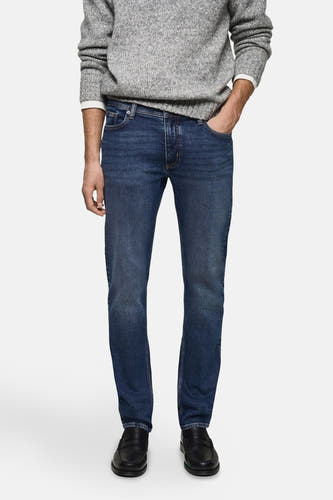 Jean slim fit en denim bleu moyen de MANGO, porté avec un pull gris et des loafers noirs.
