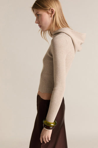 Vrouw in geribbelde beige hoodie en bruine rok, zijwaarts gericht.