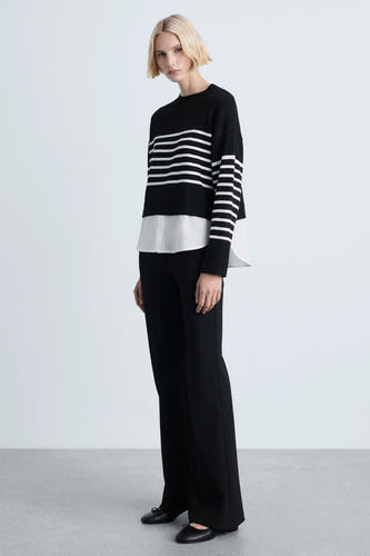 Pull noir avec col rond de MANGO, superposé à une chemise blanche et associé à un pantalon noir et des chaussures plates.