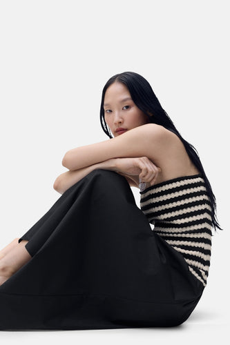 Model in gestreepte top en zwarte rok, zittend op witte achtergrond.
