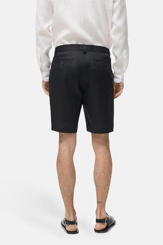 Short noir de MANGO, dos visible, associé à une chemise blanche en lin et des sandales noires.