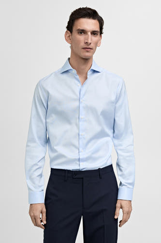 Chemise bleu clair à manches longues de MANGO, avec un col ouvert et des boutons blancs, portée sur un pantalon bleu foncé.