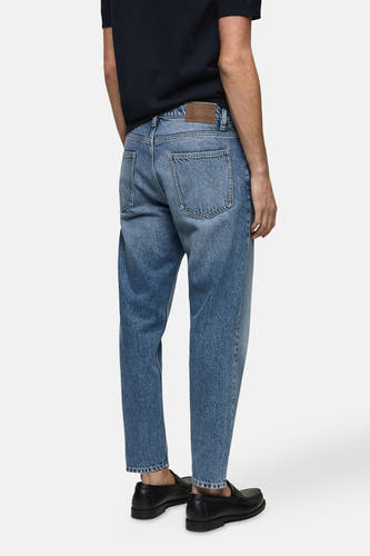 Jean tapered en denim bleu moyen de MANGO, vue arrière, avec étiquette en cuir brun et loafers noirs.