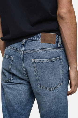 Boutons de fermeture à glissière sur des jeans en denim bleu moyen de MANGO, avec des poches arrière et une étiquette en cuir brun.