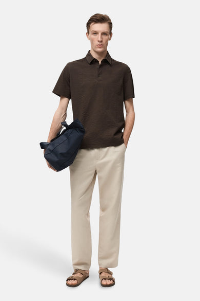 Polo marron à manches courtes de MANGO, porté avec un pantalon beige et des sandales marrons, avec une sacoche bleue.
