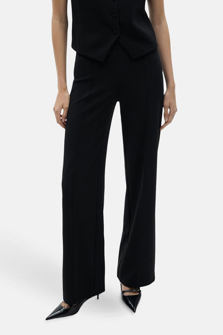 Pantalon noir de MANGO, avec un modèle qui descend et une couture centrale à l'avant, associé à des escarpins noirs et à une veste coordonnée.