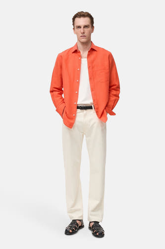 Chemise orange à manches longues de MANGO, portée sur un t-shirt blanc, avec des pantalons crème et des sandales noires.