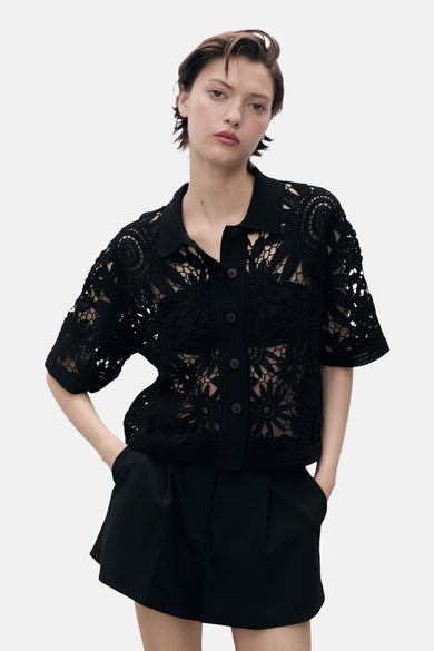Gilet noir de MANGO avec des motifs floraux ouverts, des manches courtes et une fermeture à boutons, porté avec des short noirs.
