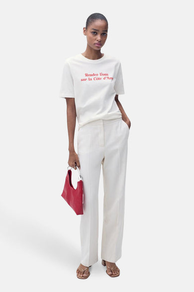 T-shirt blanc à manches courtes de MANGO, avec imprimé texte rouge et associé à un pantalon blanc et une poche main.