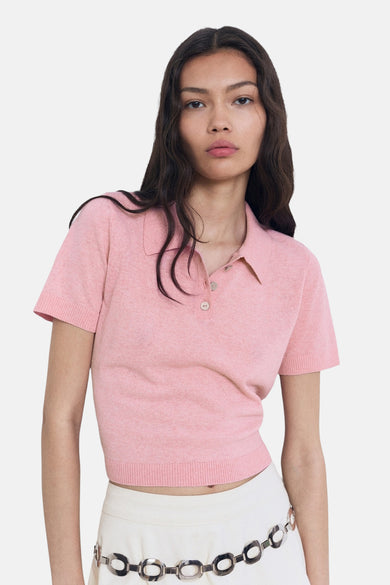 Polo rose à manches courtes de MANGO, avec une longueur courte, porté avec une jupe blanche et des sandales.