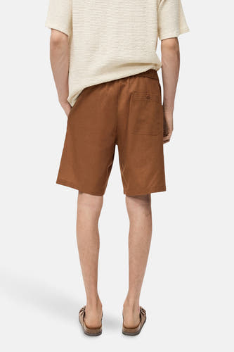 Short brun de MANGO, avec des poches à l'arrière, porté avec un t-shirt crème et des sandales brunes.