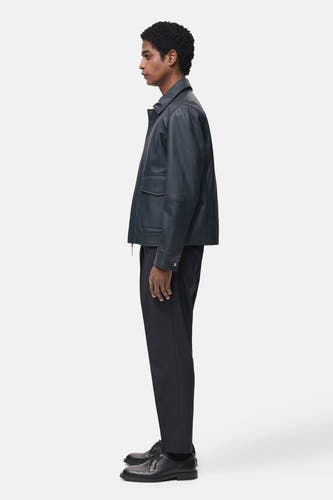 Pantalon de costume bleu MANGO, avec un motif uni, porté avec une veste en cuir et des chaussures noires, en vue latérale.