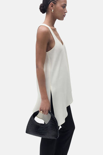 Blouse écru sans manches de MANGO, avec col V et épaules dégagées, associée à un pantalon noir et une pochette tressée noire.
